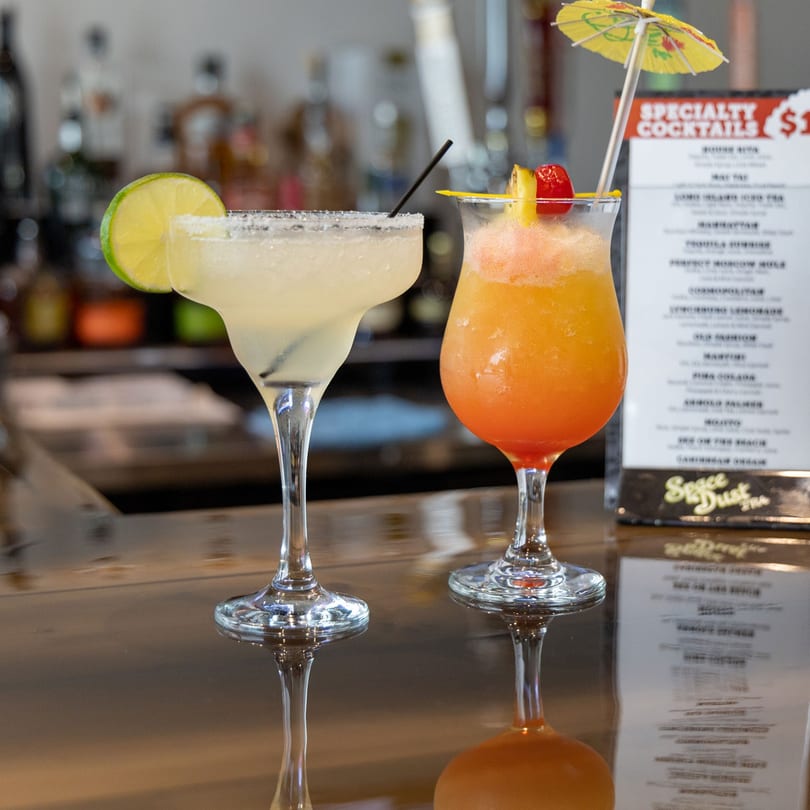 Two colorful cocktails, a margarita and a tropical drink, on a bar with a menu in the background.