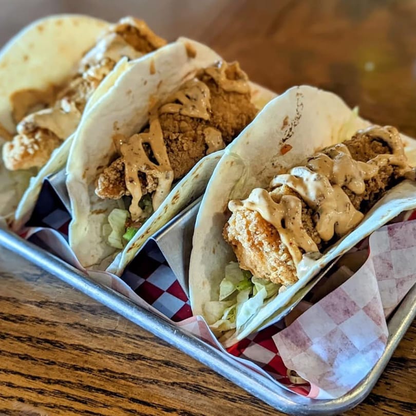Three crispy fried fish tacos with lettuce and creamy sauce in a metal tray.