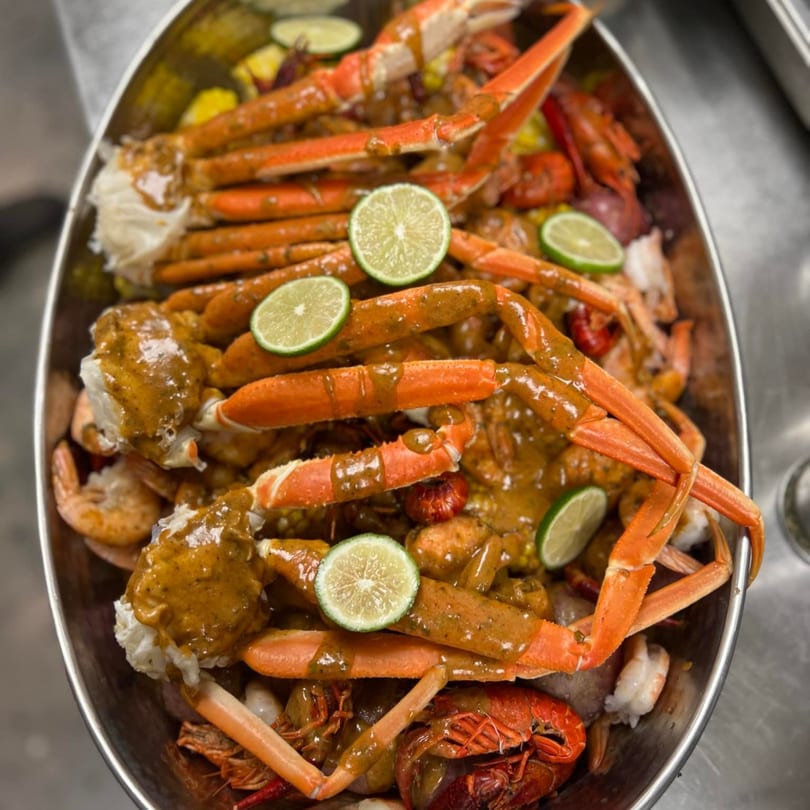 A large metal bowl filled with snow crab legs, shrimp, crawfish, and corn, garnished with lime slices.