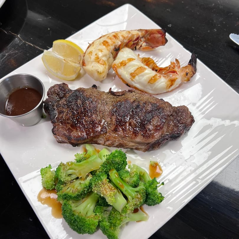Grilled steak and two large shrimp with broccoli, lemon, and dipping sauce on a white plate.