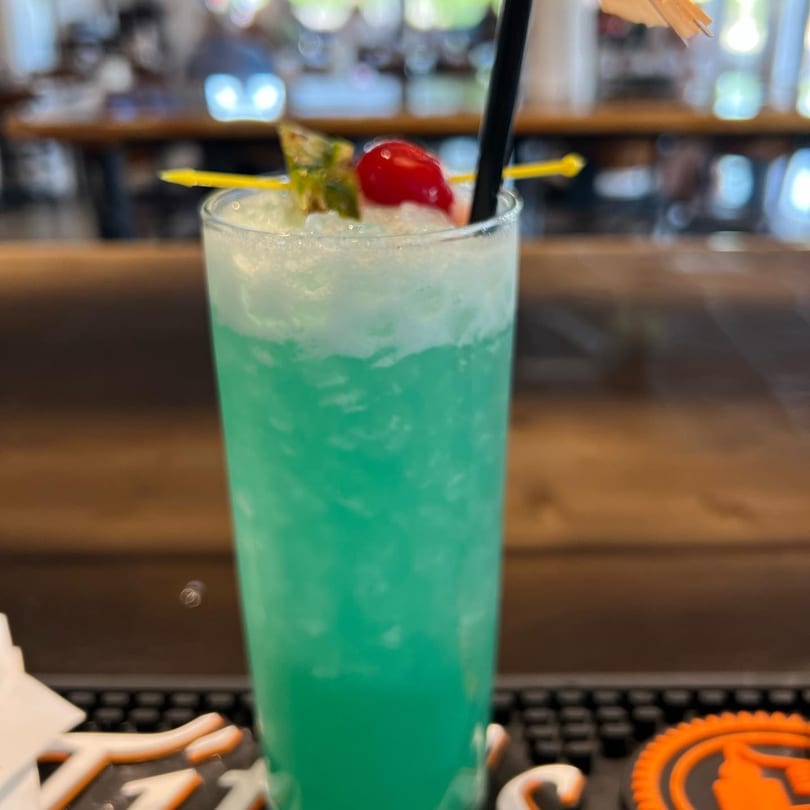A vibrant blue cocktail with a cherry and pineapple garnish sits on a bar.
